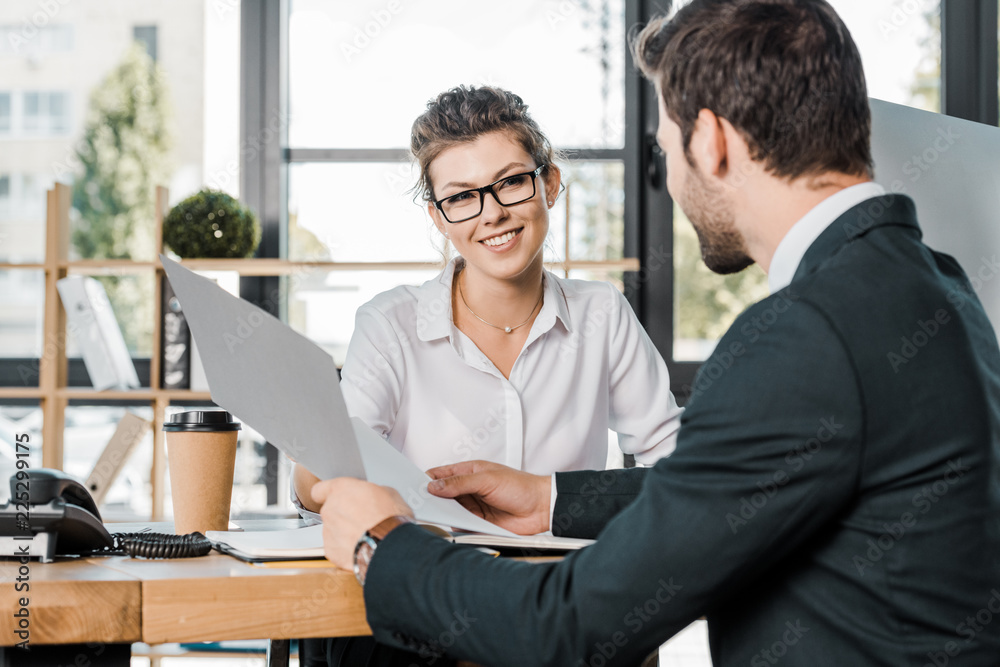 smiling businesswoman and business partner with papers discussing work at workplace in office smiling businesswoman and business partner with papers discussing work at workplace in office