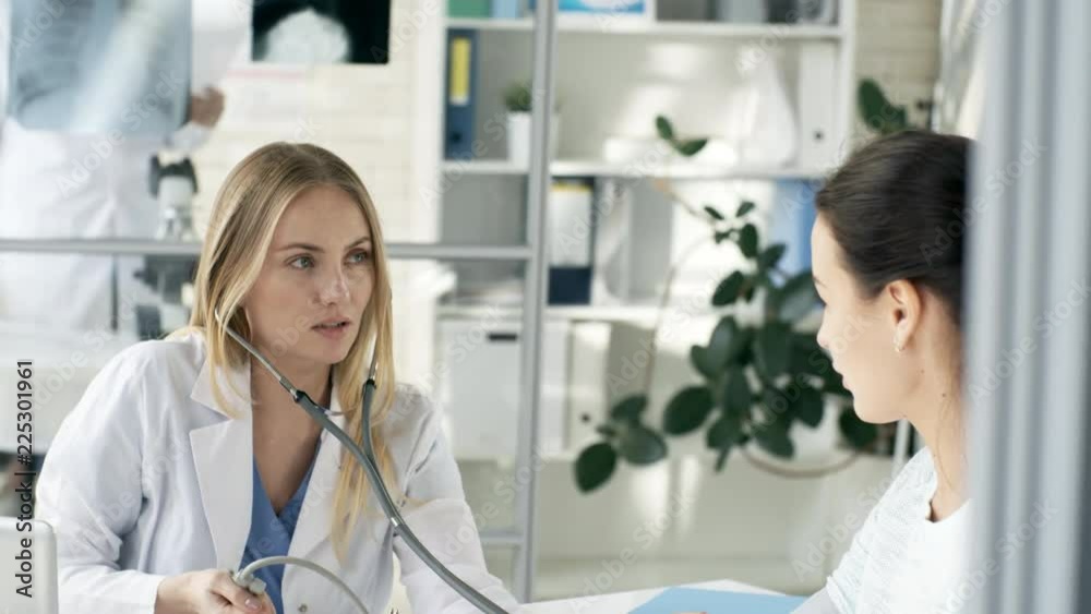 Tilt down shot of young Caucasian female doctor in lab coat smiling and talking to patient while checking blood pressure with sphygmomanometer Tilt down shot of young Caucasian female doctor in lab coat smiling and talking to patient while checking blood pressure with sphygmomanometer