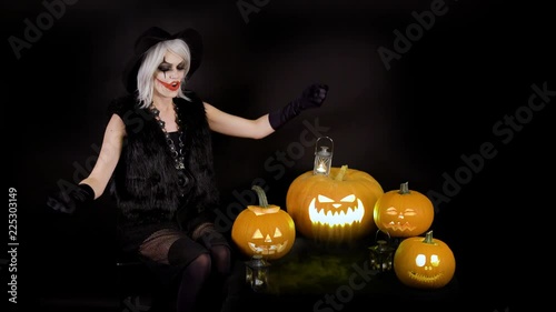 Scary beautiful girl witch laughs, taunts, gloats, celebrates halloween with funny glowing burning pumpkins in smoke. Woman with gray hair conjures, casts a spell