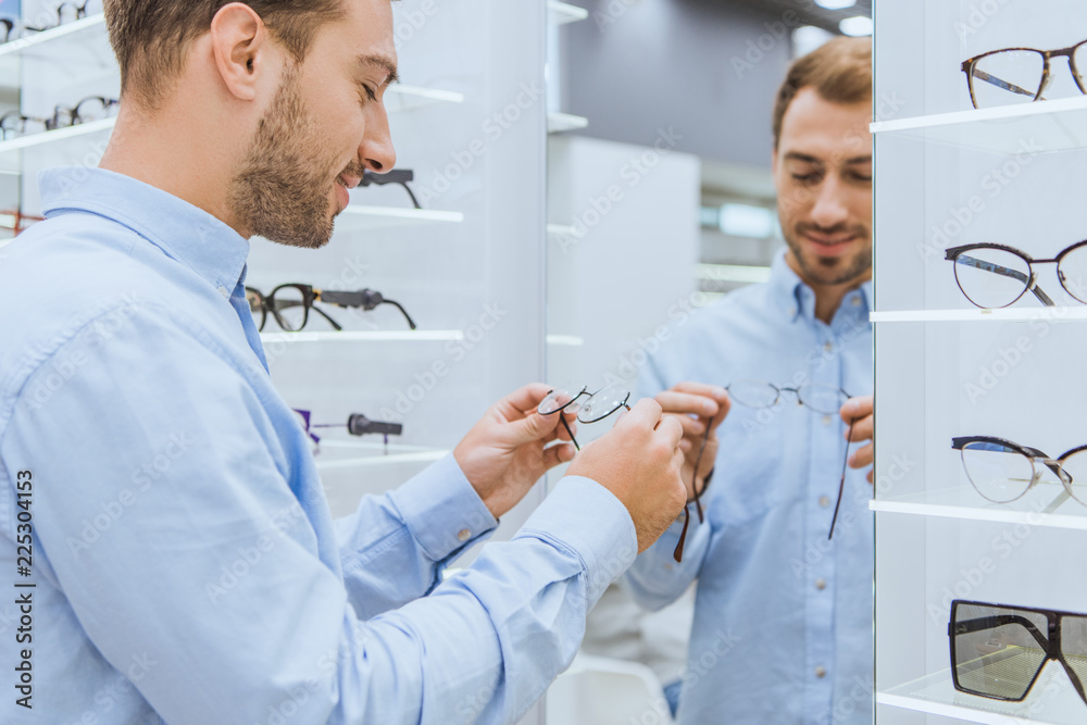 Obraz premium smiling man choosing eyeglasses in front of mirror in ophthalmic shop