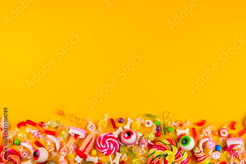 Assorted teeth & eyeball shaped candy spread on yellow background, jelly spider, gummy worms, sugar bones, round lollipop and other mixed candy, bloody finger. Top view, copy space, close up, flat lay