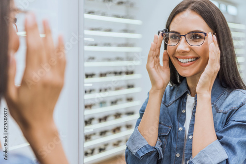 partial view of smiling woman choosing eyeglasses and looking at mirror in optica
