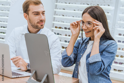 happy woman choosing eyeglasses and looking at mirror while male oculist standing near with laptop in optics