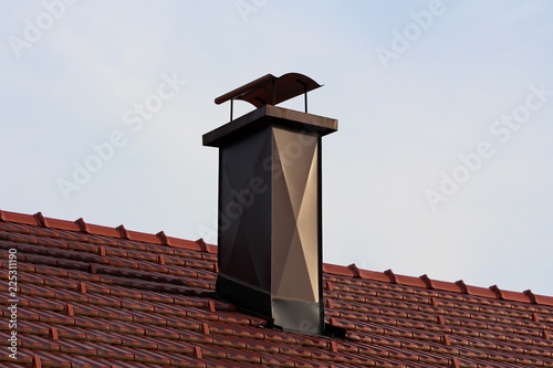Modern house chimney with brown metal protection on all sides and wavy shield on top surrounded with new red roof tiles at sunset
