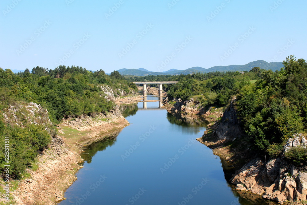 River with low summer water levels and visible rocks on river banks flowing towards small concrete bridge surrounded with lush green forest vegetation, mountains and clear blue sky in background on wa