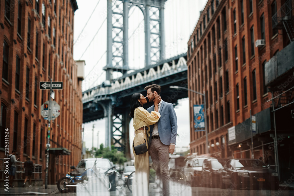 Love story in New York. Gorgeous couple of American man with beard and tender Eastern woman hug each other before the cityscape of Brooklyn bridge somewhere in New York