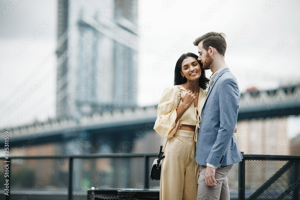 Love story in New York. Gorgeous couple of American man with beard and tender Eastern woman hug each other before the cityscape of Brooklyn bridge somewhere in New York