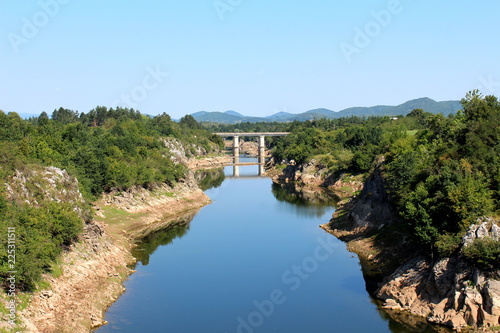 River with low summer water levels and visible rocks on river banks flowing towards small concrete bridge surrounded with lush green forest vegetation  mountains and clear blue sky in background on wa