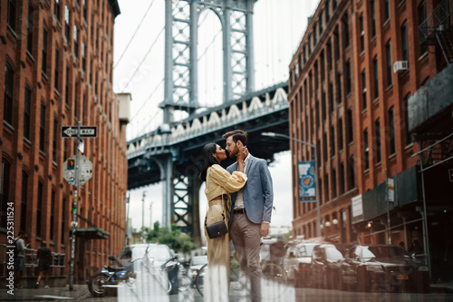 Love story in New York. Gorgeous couple of American man with beard and tender Eastern woman hug each other before the cityscape of Brooklyn bridge somewhere in New York © IVASHstudio