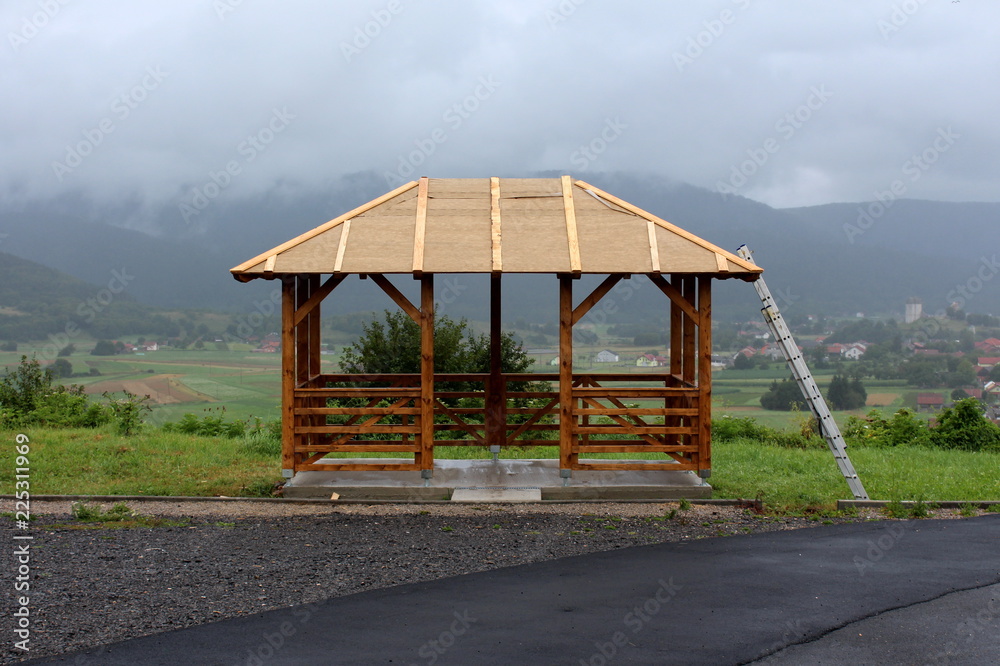 Wooden gazebo mounted on concrete foundation with roof still under construction and aluminium ladders next to it situated next to paved road on top of small hill overlooking small town and mountains i Wooden gazebo mounted on concrete foundation with roof still under construction and aluminium ladders next to it situated next to paved road on top of small hill overlooking small town and mountains i