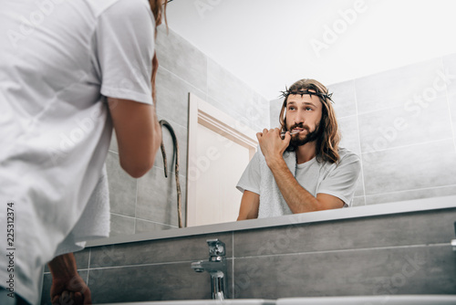 partial view of Jesus with towel over shoulder brushing teeth and looking at own reflection in bathroom