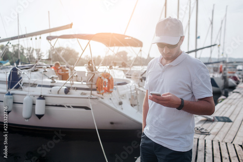 Young man stands on pier. He holds phone in hands and look at it. There are yachts behind him from one side of pier.