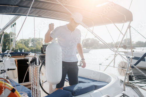 Serious sailor stands on his yacht and looks down. He holds rope with big plastic bottle. Young man is calm and peaceful. Sun goes down.