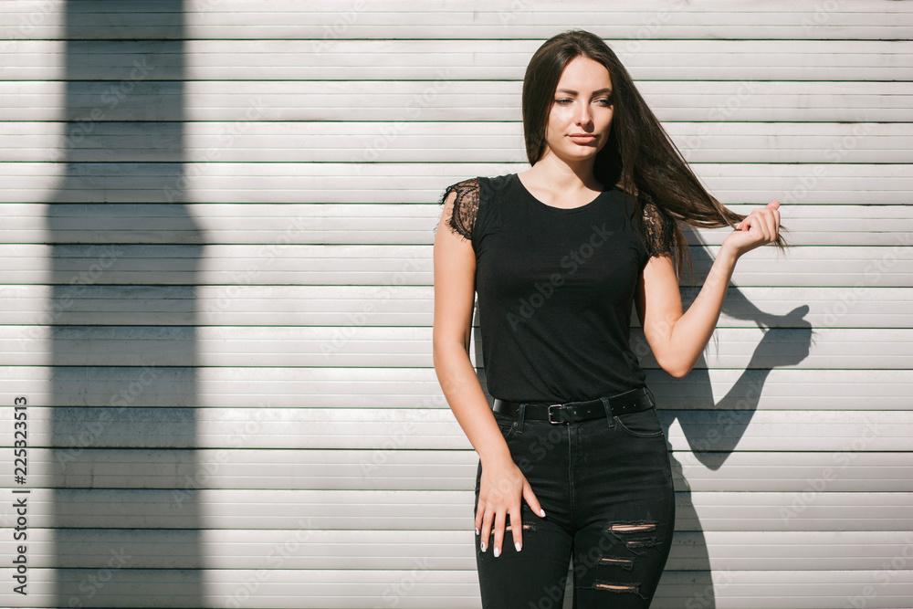 A brunette female with long hair wearing a blank black t-shirt i while standing on a concrete wall background on a street. Empty space for text or design. A brunette female with long hair wearing a blank black t-shirt i while standing on a concrete wall background on a street. Empty space for text or design.