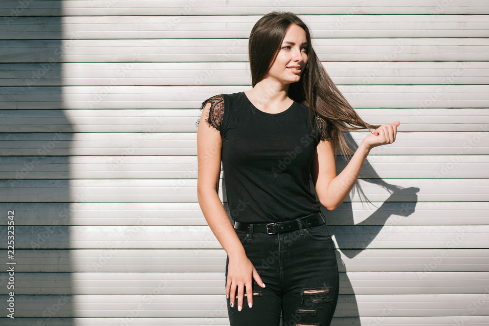Fototapeta premium A brunette female with long hair wearing a blank black t-shirt i while standing on a concrete wall background on a street. Empty space for text or design.