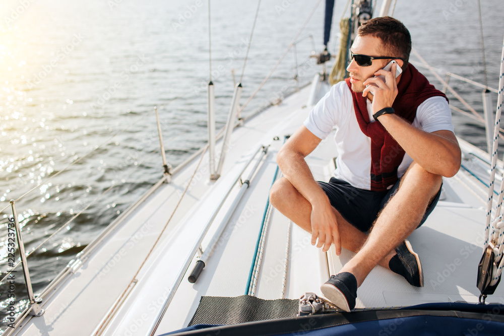 Young man sits on yacht board and look to left. He talks on phone. Guy sits with legs crossed. He wears sunglasses. Young man is busy.