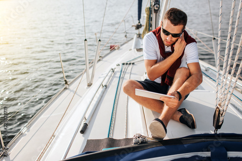 Busy young man sits on board of yacht and talks on phone. Also he holds and looks at tablet. Young man sits with legs crossed. He wears sunglasses.