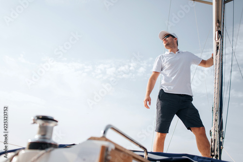 Young man stands on yacht board and looks forward. He holds on mast with hand. Young man poses. He wears white shirt and black shorts.