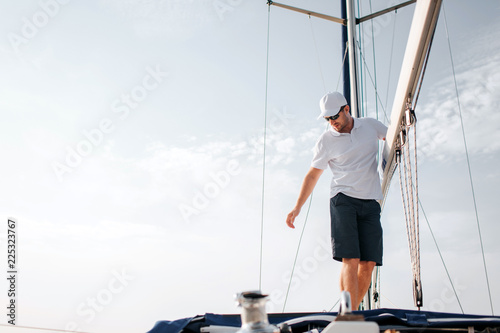 Handsome and confident man stands on yacht board and holds on big mast. He looks down and reaches with hand. Young man is serious and concentrated. He poses.