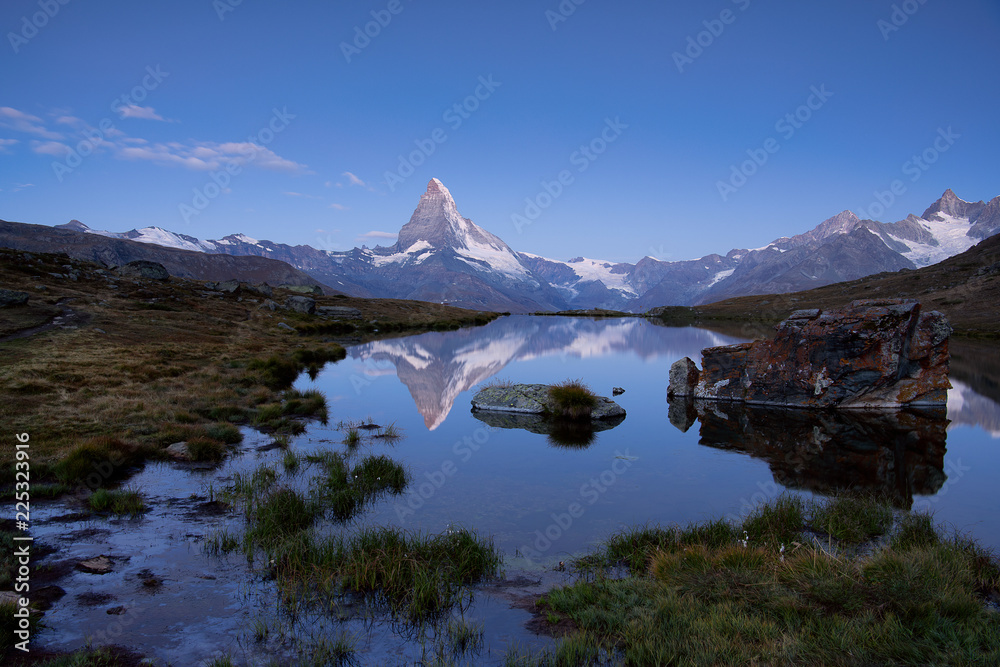 Unspoilt Matterhorn and ridge with snow fields in blue hour and cold blue sky reflected on water Stellisee with stones and grass
