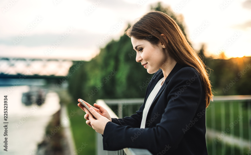 Fototapeta premium A young businesswoman standing on the river bank, using smartphone.