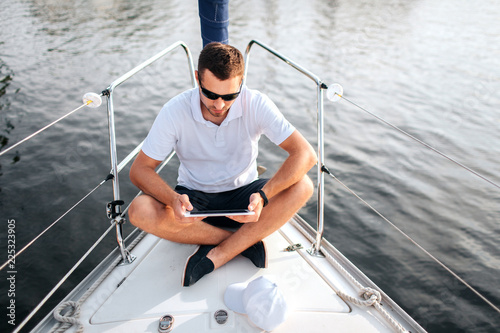 Young man sits with his legs crossed and hold tablet in hands. He is calm and concentrated. Screen is black. Guy sits on yacht bow. He wears sunglasses. © estradaanton