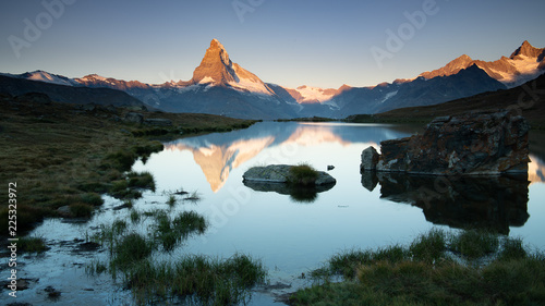 Sunrise at unspoilt Matterhorn with warm soft light on ridge and snow fields, clear blue sky, reflected on water Stellisee with stones and grass