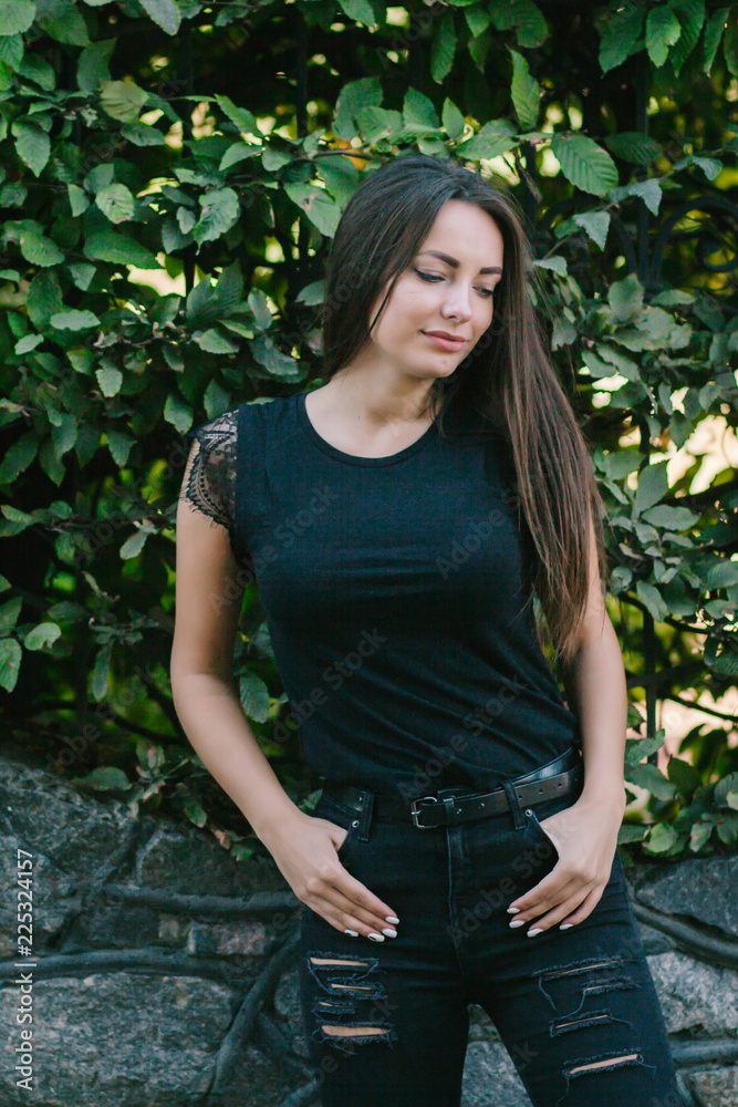 A brunette female with long hair wearing a blank black t-shirt i while standing on a concrete wall background on a street. Empty space for text or design. A brunette female with long hair wearing a blank black t-shirt i while standing on a concrete wall background on a street. Empty space for text or design.