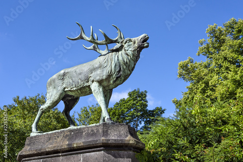 Fotografie Big bronze statue of an antlered belling stag in a green park with trees and blu