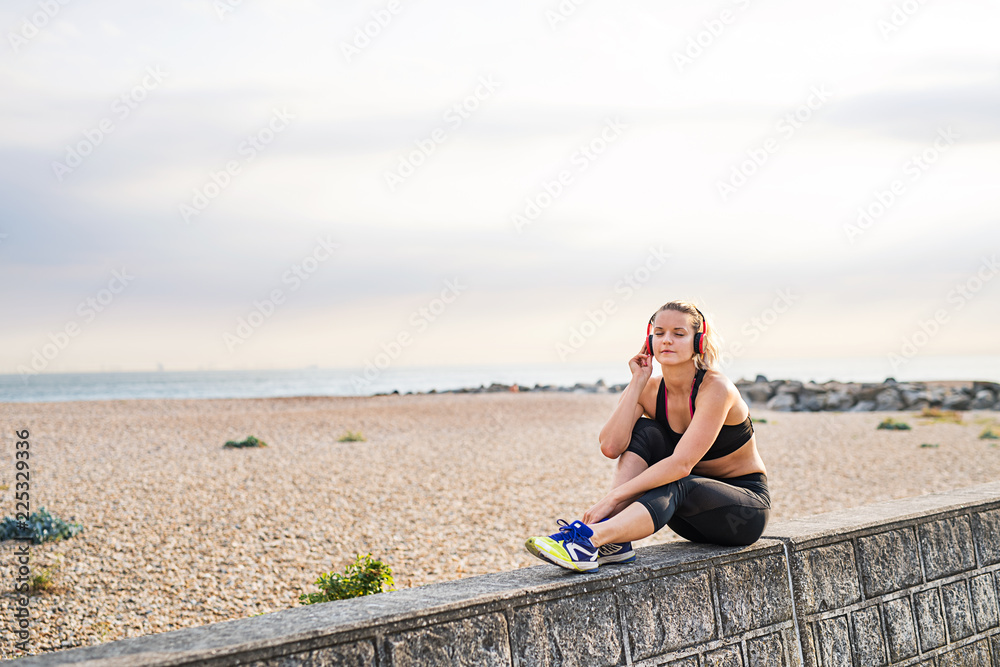 A young woman runner with headphones resting outside by the sea.