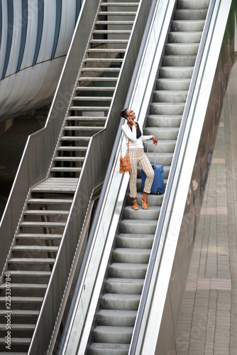 happy african american woman standing on escalator with travel bags and mobile phone