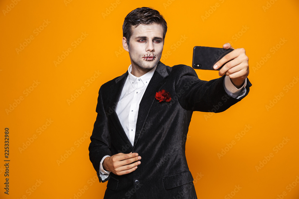 Photo of terrible dead man on halloween wearing classical suit and creepy makeup taking selfie on mobile phone, isolated over yellow background Photo of terrible dead man on halloween wearing classical suit and creepy makeup taking selfie on mobile phone, isolated over yellow background