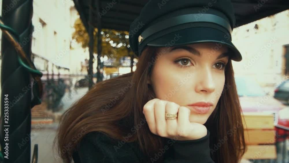 Charming fashion model in black cap. Portrait of glamorous young woman relaxing outdoors. Close up face of elegant woman with beautiful eyes. Fashionable girl posing in urban landscape at daylight