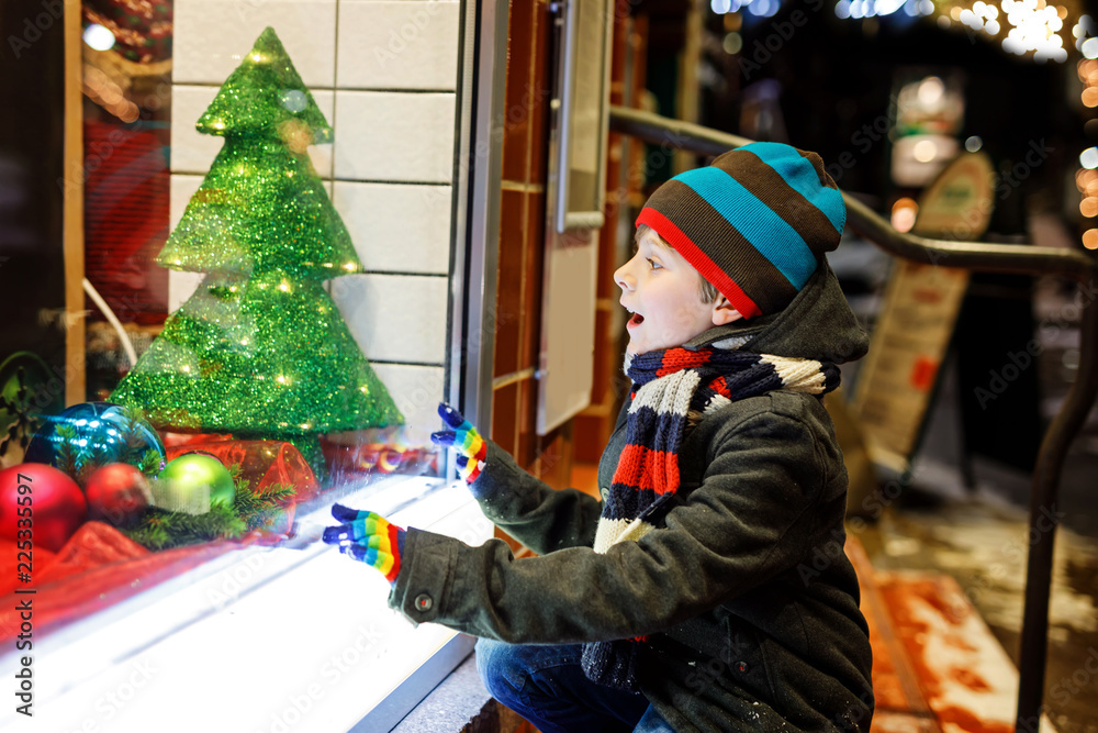 Funny happy child in fashion winter clothes making window shopping decorated with gifts, xmas tree