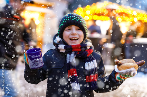 Little cute kid boy eating German sausage and drinking hot children punch on Christmas market photo