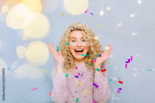 Happy blond curly hipster girl enjoying and celebrating on a grey background. Blows up multicolored confetti. Photo taken through the garland light. Leisure, winter, holidays, people concept.