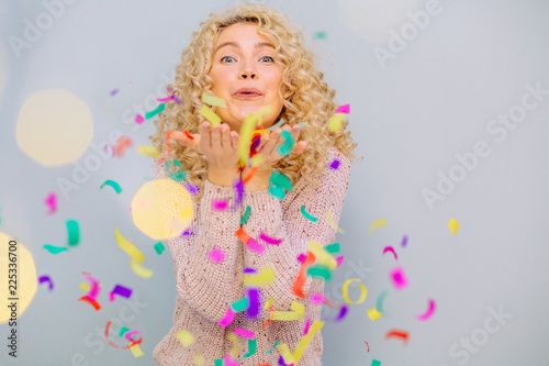 Happy blond curly hipster girl celebrating on a grey background. Blows up multicolored confetti.