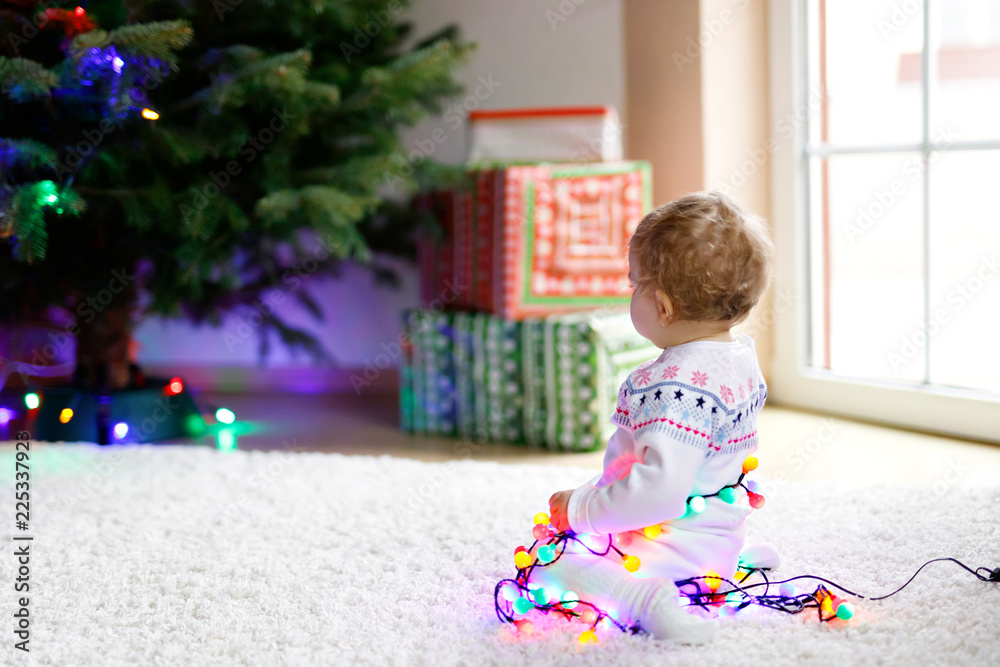 Adorable baby girl holding colorful lights garland in cute hands. Little child in festive clothes decorating Christmas tree