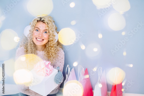 A curly blond girl smiles and happy while shopping in the stores with sales at winter time. Curly woman holds present box and paper bags. Concept of: shopping, holidays, happiness, presents. photo