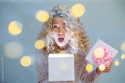 Schocked curly blonde woman in knitted sweater opening present box with pink bow indoor over gray wall background.Photo taken through the garland. Leisure, hygge, people concept. Close up photo