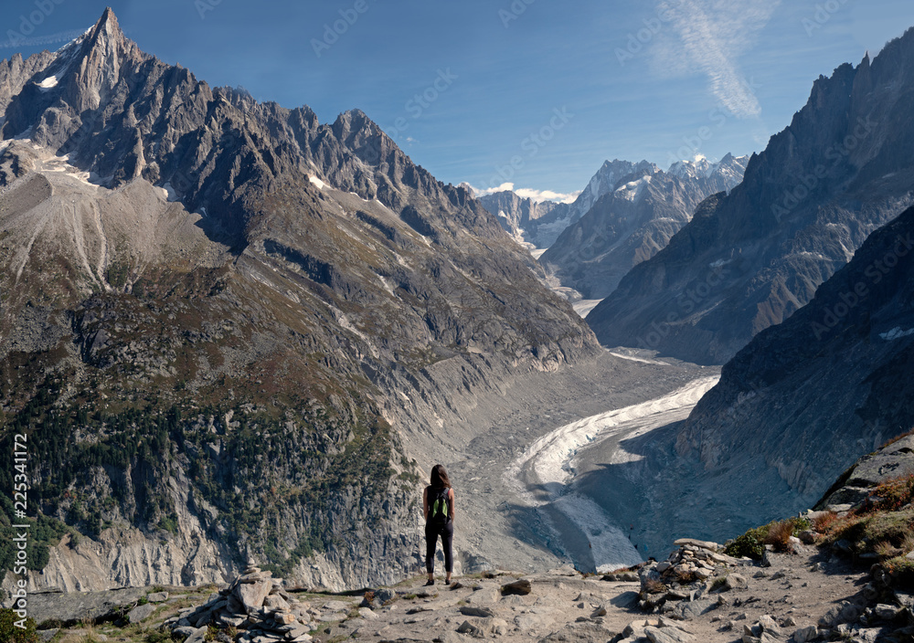 A lone female Hiker looks out over a  glacier valley in the region of Chamonix in the alps of France. High alpine snow covered mountains surround the foreground rocks and alpine terrain.