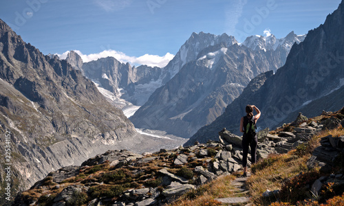 A lone female Hiker looks out over a glacier valley in the region of Chamonix in the alps of France. High alpine snow covered mountains surround the foreground rocks and alpine terrain.