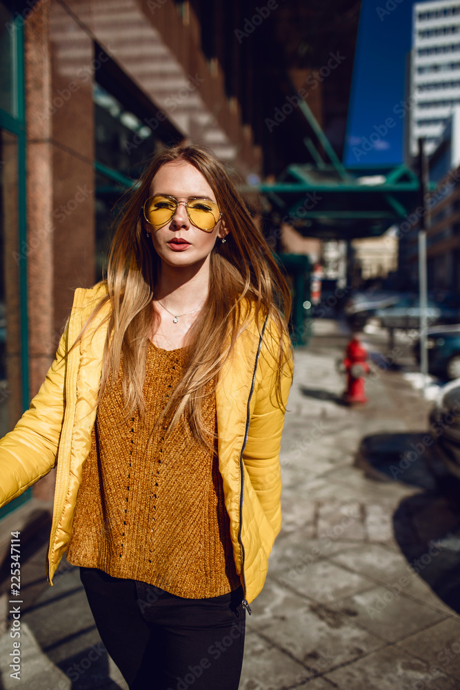 A young European woman, traveling, with long blond hair, wearing a yellow jacket, yellow sunglasses walking down the city center street, street shooting. Even light. A young European woman, traveling, with long blond hair, wearing a yellow jacket, yellow sunglasses walking down the city center street, street shooting. Even light.