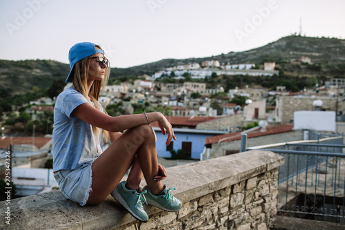 Beautiful young sexy hipster girl posing and smiling near urban blue wall background in yellow dress, shorts, shirt, sneakers, sunglasses.