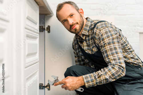 handsome plumber holding adjustable wrench near kitchen cabinet and looking at camera