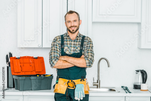 smiling handsome plumber standing with crossed arms and looking at camera in kitchen