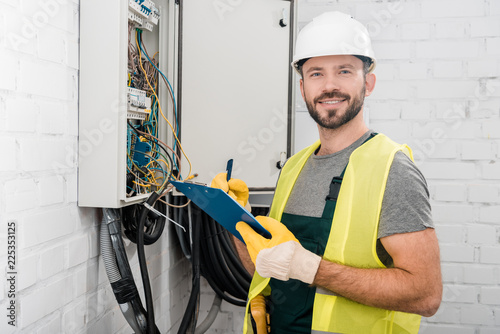 smiling handsome electrician holding clipboard near electrical box in corridor and looking at camera