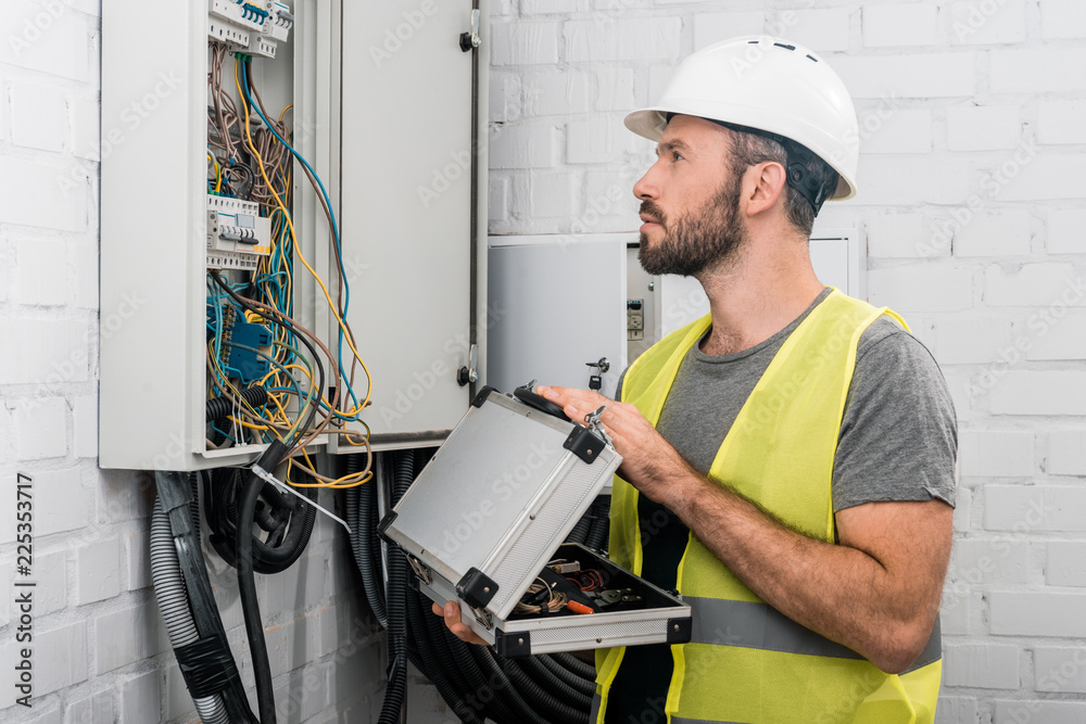 side view of handsome electrician holding toolbox and looking at electrical box in corridor side view of handsome electrician holding toolbox and looking at electrical box in corridor