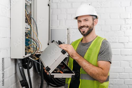 smiling electrician holding toolbox near electrical box in corridor and looking at camera photo