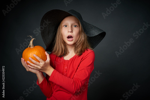 Closeup of surprised girl in black hat holding pumpkin over dark background